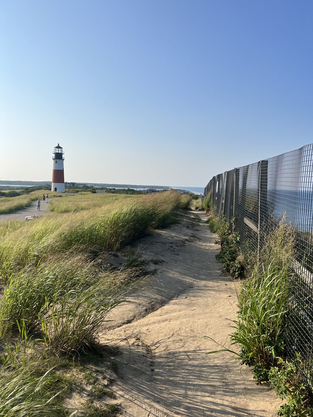 A sand path along the fence at the east-end bluffs, the lighthouse beyond and the Atlantic to the right.