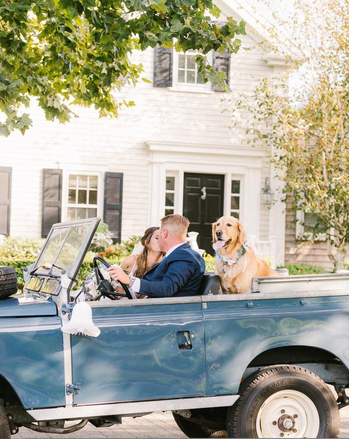 A couple kissing in the front of the Land Rover, their golden retriever riding in the back.