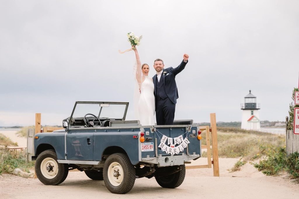 A just-married couple standing in the back of a classic Land Rover on the beach, Brant Point Light behind them.